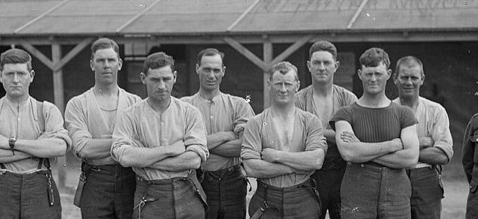 Eight young, strong soldiers wearing casual clothes stand in a yard with their arms crossed.