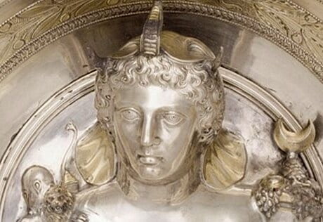 A woman's face sculpted in silver. She wears an elephant headdress, she has wavy hair, a forehead that slants backwards, curved eyebrows, big eyes, an aquiline nose, small mouth, prominent rounded chin. The work is Roman or Greek.