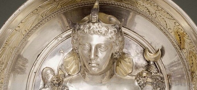 A woman's face sculpted in silver. She wears an elephant headdress, she has wavy hair, a forehead that slants backwards, curved eyebrows, big eyes, an aquiline nose, small mouth, prominent rounded chin. The work is Roman or Greek.